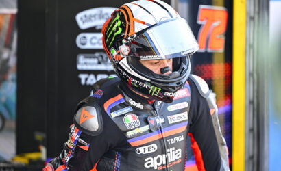 Motorsport rider in a black/orange Aprilia racing suit and full-face helmet, standing in the pit area with sponsor logos visible on the suit and helmet.