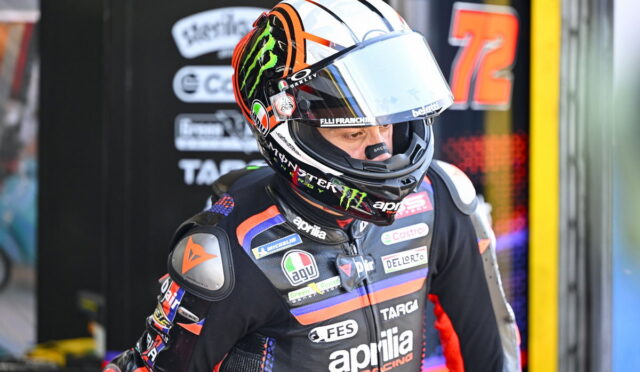 Motorsport rider in a black/orange Aprilia racing suit and full-face helmet, standing in the pit area with sponsor logos visible on the suit and helmet.