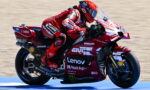 Rider in red race suit leaning into a red Ducati MotoGP bike with sponsor logos (Lenovo, Shell, Monster) on a racetrack surface