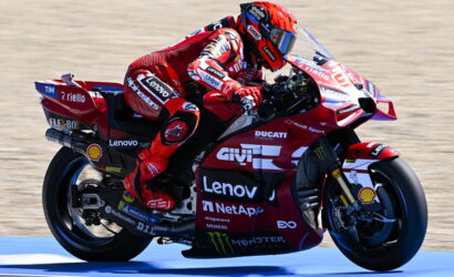 Rider in red race suit leaning into a red Ducati MotoGP bike with sponsor logos (Lenovo, Shell, Monster) on a racetrack surface