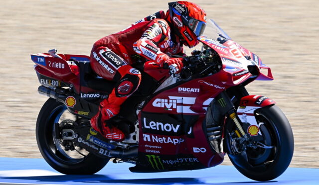 Rider in red race suit leaning into a red Ducati MotoGP bike with sponsor logos (Lenovo, Shell, Monster) on a racetrack surface