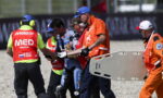 Emergency responders and a motorcycle racer escorting across a gravel track, with a medic wearing a bright red 'MEDICAL' vest and a team member holding a stretcher nearby.