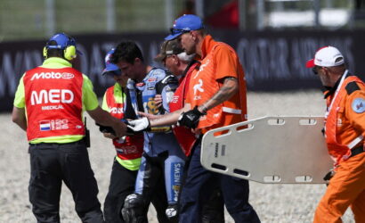 Emergency responders and a motorcycle racer escorting across a gravel track, with a medic wearing a bright red 'MEDICAL' vest and a team member holding a stretcher nearby.