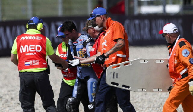 Emergency responders and a motorcycle racer escorting across a gravel track, with a medic wearing a bright red 'MEDICAL' vest and a team member holding a stretcher nearby.