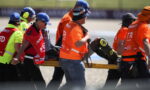Emergency responders in bright orange vests carry a patient on a yellow stretcher across a racing track.