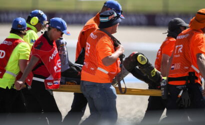 Emergency responders in bright orange vests carry a patient on a yellow stretcher across a racing track.