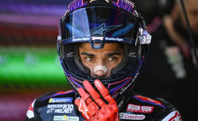 Motocross racer in a purple helmet with red gloves, hand near the face shield, looking ahead.