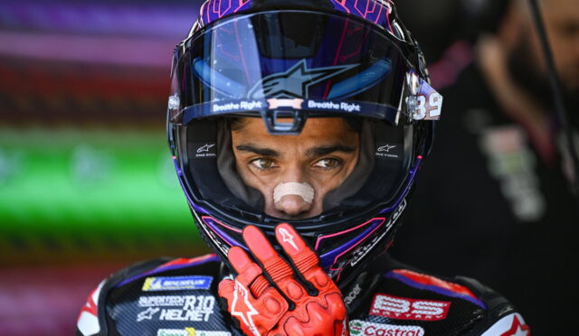 Motocross racer in a purple helmet with red gloves, hand near the face shield, looking ahead.