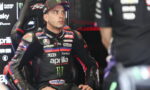 MotoGP rider in a black Aprilia racing suit seated in the pit, looking to the left.
