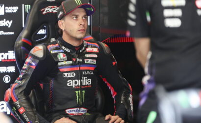 MotoGP rider in a black Aprilia racing suit seated in the pit, looking to the left.