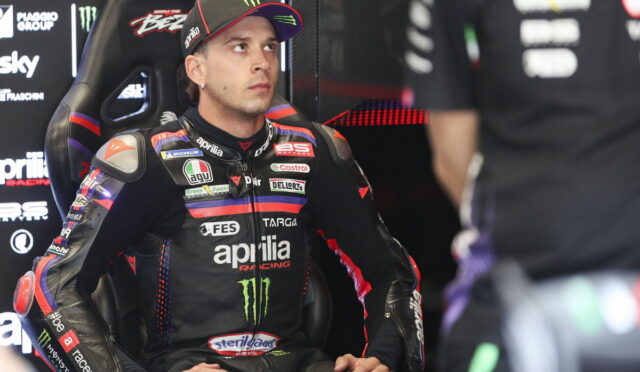 MotoGP rider in a black Aprilia racing suit seated in the pit, looking to the left.