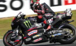 MotoGP rider in black and red gear leans into a turn on a sponsor-covered racing bike with Monster Energy and Castrol logos.