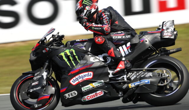 MotoGP rider in black and red gear leans into a turn on a sponsor-covered racing bike with Monster Energy and Castrol logos.