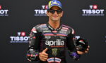 MotoGP rider in an Aprilia racing suit gives a thumbs-up while holding a helmet, in front of a Tissot backdrop.