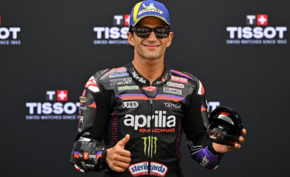 MotoGP rider in an Aprilia racing suit gives a thumbs-up while holding a helmet, in front of a Tissot backdrop.