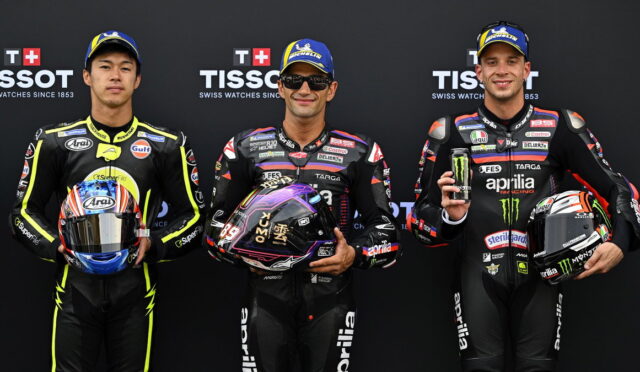 Three motorcycle racers in full leathers stand side by side, each holding a helmet, posing for a photo.