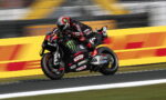 Motorcycle racer in black and red gear leaning into a turn on a race track with sponsor logos visible on the bike.