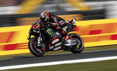 Motorcycle racer in black and red gear leaning into a turn on a race track with sponsor logos visible on the bike.