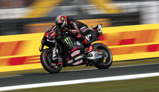 Motorcycle racer in black and red gear leaning into a turn on a race track with sponsor logos visible on the bike.
