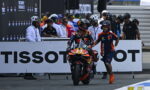 MotoGP rider on an orange-red racing bike with Red Bull livery, helmet on, being guided by a crew member in blue and orange at a pit wall behind a TISSOT banner.