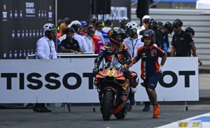 MotoGP rider on an orange-red racing bike with Red Bull livery, helmet on, being guided by a crew member in blue and orange at a pit wall behind a TISSOT banner.