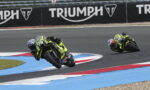 Two motorcyclists in neon-yellow gear lean into a sharp turn on a racetrack, Triumph banners along the fence in the background.