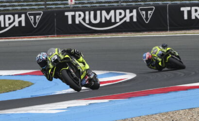 Two motorcyclists in neon-yellow gear lean into a sharp turn on a racetrack, Triumph banners along the fence in the background.