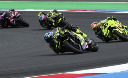 Motorcycle racers in neon yellow gear lean into a sharp track bend during a race, close behind each other on a dark asphalt circuit.