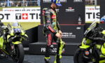 Motorsport rider in a neon-yellow and black racing suit stands between two yellow racing motorcycles on a podium, with sponsor banners in the background.