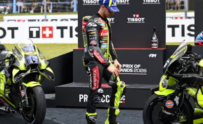 Motorsport rider in a neon-yellow and black racing suit stands between two yellow racing motorcycles on a podium, with sponsor banners in the background.
