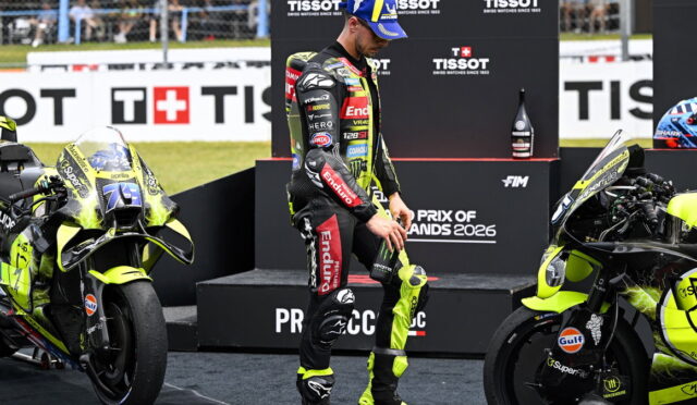Motorsport rider in a neon-yellow and black racing suit stands between two yellow racing motorcycles on a podium, with sponsor banners in the background.