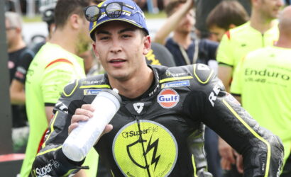 Motorcycle racer in black and neon-yellow suit drinks from a water bottle, surrounded by helpers and spectators during a race event.