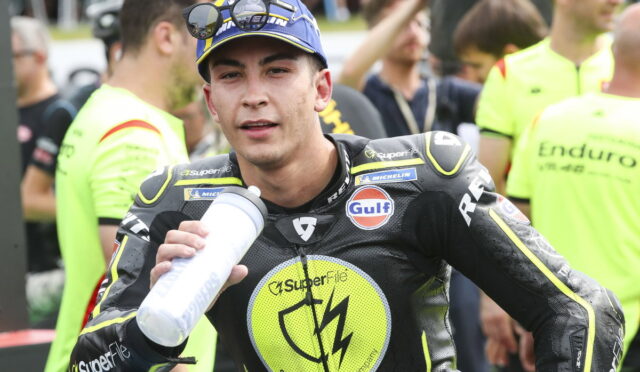 Motorcycle racer in black and neon-yellow suit drinks from a water bottle, surrounded by helpers and spectators during a race event.