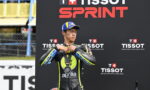 MotoGP rider in a black and yellow Aprilia racing suit adjusts his medal on the podium at the Tissot Sprint event, backdrop with TISSOT branding.
