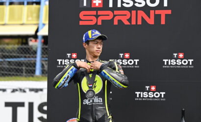 MotoGP rider in a black and yellow Aprilia racing suit adjusts his medal on the podium at the Tissot Sprint event, backdrop with TISSOT branding.