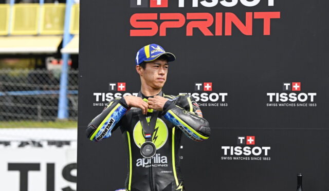 MotoGP rider in a black and yellow Aprilia racing suit adjusts his medal on the podium at the Tissot Sprint event, backdrop with TISSOT branding.