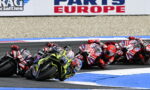 Group of motorcycle racers leaning into a sharp turn on a paved race track, red and black bikes leading the pack.