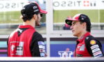 Two men in red and black racing uniforms chat in a paddock, sponsor logos visible on their jackets.