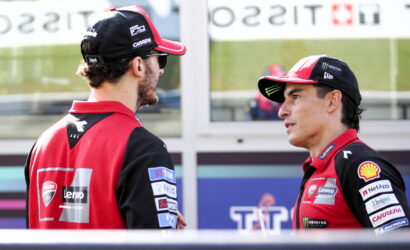 Two men in red and black racing uniforms chat in a paddock, sponsor logos visible on their jackets.