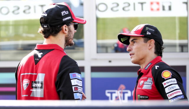 Two men in red and black racing uniforms chat in a paddock, sponsor logos visible on their jackets.