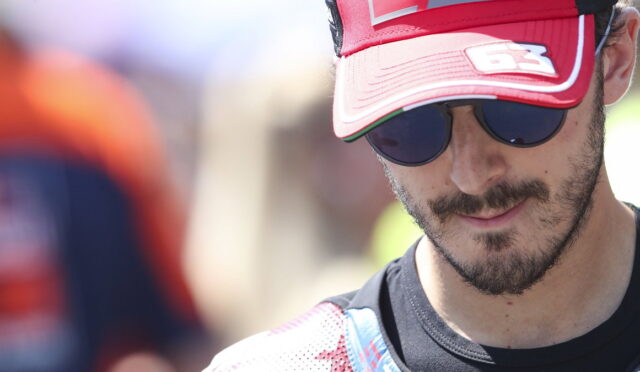 Man wearing a red cap and dark sunglasses, partial face visible outdoors, slight smile on lips.