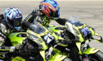 Two riders on neon-yellow Aprilia race bikes lean into a turn on a race track, competing closely beside each other to the left.