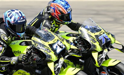Two riders on neon-yellow Aprilia race bikes lean into a turn on a race track, competing closely beside each other to the left.