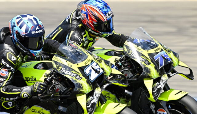 Two riders on neon-yellow Aprilia race bikes lean into a turn on a race track, competing closely beside each other to the left.