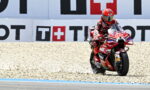 Moto racing rider in red suit on a red motorcycle (number 93) speeding along a gravel run-off by advertising boards at the track edge.