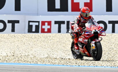Moto racing rider in red suit on a red motorcycle (number 93) speeding along a gravel run-off by advertising boards at the track edge.