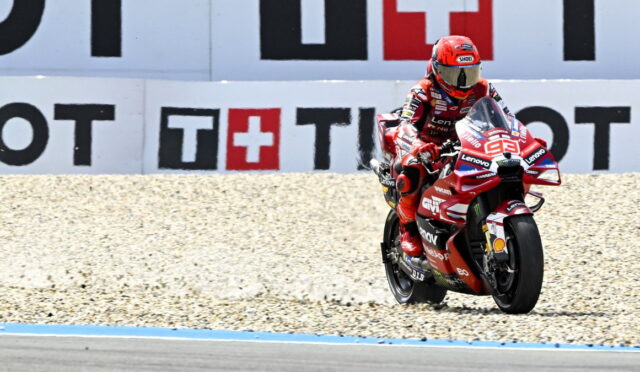 Moto racing rider in red suit on a red motorcycle (number 93) speeding along a gravel run-off by advertising boards at the track edge.