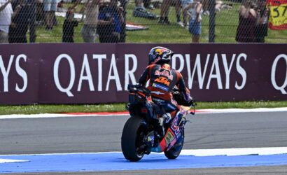 MotoGP rider in Red Bull KTM gear exits a corner on a race track; blue runoff area and spectators behind a fence with Qatar Airways advertising