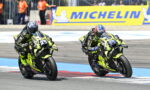 Two neon-yellow race motorcycles lean into a sharp turn on a race track with riders in matching black and neon-yellow gear and a Michelin banner in the background.