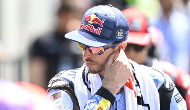 Motorsport rider in a blue Red Bull cap and mirrored sunglasses, touching his ear with a towel draped over his shoulders.